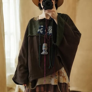 May include: A brown, long-sleeved top with a patchwork design. The top features a green yoke and sleeves, with a blue and white patterned patch. A decorative pendant hangs from the neckline. The style is bohemian.