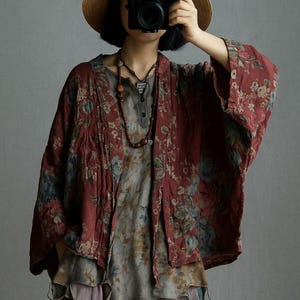 May include: A red floral print kimono-style jacket with a loose fit, layered over a gray and floral print top. The jacket features a dark red base with blue and pink floral patterns. A necklace with wooden beads and a pendant is visible.