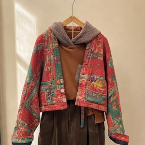 May include: A red, green, and gold patterned jacket with a brown corduroy bottom. The jacket has a gray hood and a brown shirt underneath. The jacket has three buttons and a pocket. The jacket is hanging on a wooden hanger.