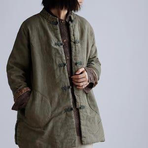 Può includere: Giacca verde oliva a maniche lunghe con collo alla coreana e chiusure a frog. La giacca è sovrapposta a un maglione marrone. Le maniche sono arrotolate, rivelando una fodera fantasia. Tasche frontali.