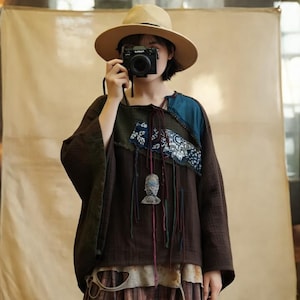 May include: A brown long-sleeve top with patchwork details in blue and green, and a decorative pendant. The top has a square neckline with string ties and is paired with a straw hat. The outfit is a bohemian style.