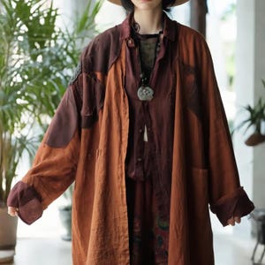 Puede incluir: Un abrigo largo y abierto en tonos marrón y burdeos. El abrigo presenta un diseño de patchwork en las mangas y la parte superior de la espalda, con bolsillos grandes. El atuendo incluye una camisa oscura y un collar con colgante floral. El estilo es casual y bohemio.