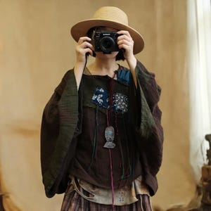 May include: A brown and green top with patchwork details, a straw hat, and a camera. The top features patches of blue and white patterned fabric and a decorative pendant. The sleeves are wide and flowing, creating a bohemian aesthetic.