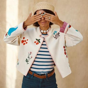 Puede incluir: Chaqueta crema bordada con corazones rojos, cerezas y diseños florales. La chaqueta tiene un ribete rosa y se usa sobre una camisa a rayas y vaqueros azules. Un sombrero de paja y un collar de margaritas completan el look.