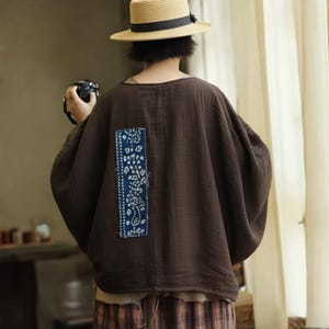 May include: A brown, oversized top with a unique blue and white floral patch on the back. The top has a relaxed fit and long sleeves. The model is wearing a straw hat with a black band.