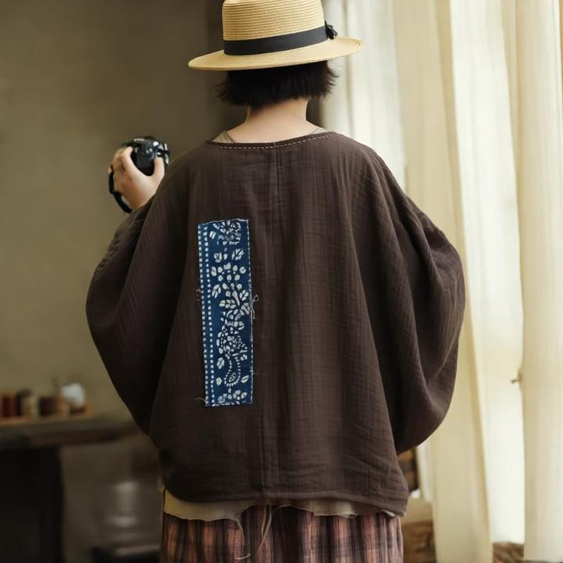 May include: A brown, oversized top with a unique blue and white floral patch on the back. The top has a relaxed fit and long sleeves. The model is wearing a straw hat with a black band.