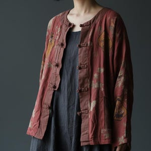 Könnte beinhalten: Rote Jacke mit Blumenmuster, Stehkragen und Knöpfen. Die Jacke wird über einem grauen Kleid getragen. Die Ärmel der Jacke sind lang.