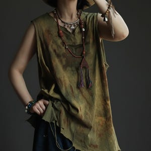 Puede incluir: Top sin mangas verde oliva con cuello en V y efecto tie-dye desgastado. El top se combina con un collar largo de cuentas y pulseras. El estilo general es bohemio.