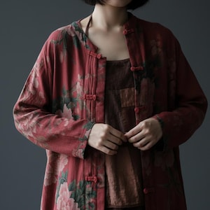 Peut inclure: Long manteau rouge à imprimé floral avec col mandarin et fermetures à brandebourgs. Le manteau présente un grand motif floral de pivoines dans des tons de rose, vert et blanc. Porté sur un haut marron.