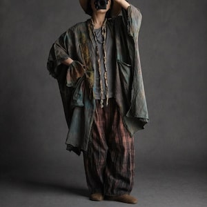 Puede incluir: Una chaqueta fluida de estilo kimono de gran tamaño en tonos verdes, azules y marrones apagados, combinada con pantalones a cuadros. La chaqueta presenta un diseño de bloques de color único y mangas anchas. El atuendo se completa con un sombrero y una bufanda larga y texturizada.