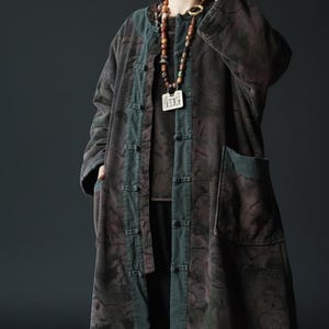 Może przedstawiać: Długi płaszcz sztruksowy w odcieniach brązu i zieleni z kwiatowym wzorem. Płaszcz ma zapięcie na guziki, kieszenie i kontrastowe zielone wykończenia. Widoczny jest długi naszyjnik z koralików z wisiorkiem.