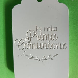 Op de afbeelding: Een witte tag met een geschulpte rand en een gat om op te hangen. De tekst "la mia Prima Comunione" is in sierlijke letters gegraveerd, met een decoratief takje aan de onderkant.