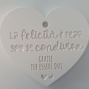 Può includere: Una placca di legno bianca a forma di cuore con un foro nella parte superiore. La placca presenta un testo italiano inciso che recita "La felicità è reale solo se condivisa. Grazie per essere qui" e due piccole forme di cuore.