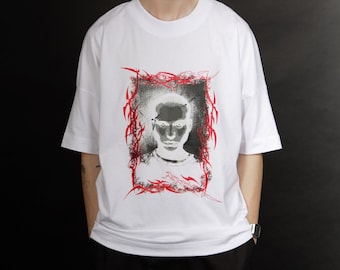 Op de afbeelding: Wit T-shirt met een grafische print van een gezicht in een rood frame. Het frame is gemaakt van gestileerde lijnen.