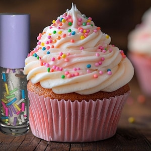 May include: A cupcake with pink icing and colourful sprinkles next to a lip gloss tube with a light purple cap. The lip gloss contains colourful, sprinkle-shaped glitter. The cupcake is in a pink paper liner.