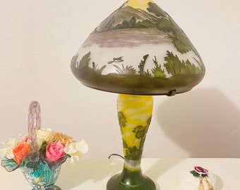 May include: A green and yellow glass table lamp with a white and green shade featuring a painted landscape scene. The shade is shaped like a mushroom cap.