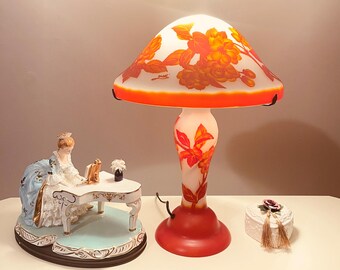 May include: A decorative table lamp with a mushroom-shaped shade, adorned with orange and yellow floral patterns. The lamp has a red base and stem. A porcelain figurine and a small box are also visible.