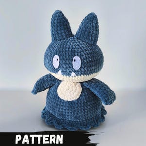 Pode incluir: Um brinquedo de pelúcia de crochê em forma de um personagem Pokemon azul e creme. O brinquedo tem olhos grandes e uma boca larga. A palavra "PATTERN" está escrita em texto preto sobre um fundo branco na parte inferior da imagem.