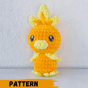 May include: A crocheted plush toy in the shape of a yellow and orange bird with a yellow beak and purple eyes. The toy is standing on a white surface. PATTERN is written in black letters on a yellow background at the bottom of the image.