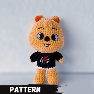 Puede incluir: Un juguete de peluche de crochet con forma de oso que lleva una camisa negra con un gráfico rojo. El oso tiene orejas naranjas y pelaje marrón.