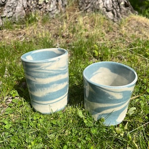 May include: Two ceramic tumblers with a blue and white marbled design. The cups are cylindrical with a slightly flared rim. They are sitting on green grass in front of a tree.