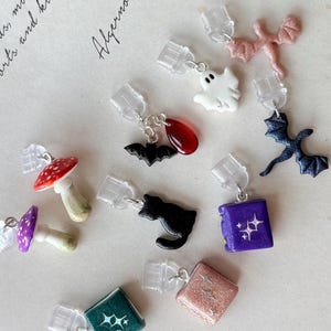 May include: A collection of small charms featuring various shapes including a white ghost, a black bat with a red drop, a pink dragon, a dark blue dragon, two mushrooms, a black cat, and three books in purple, green, and glittery peach colors.