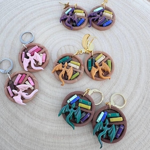 May include: A pair of earrings featuring a dragon flying over a bookshelf with colorful books. The earrings are made of polymer clay and have gold hooks.