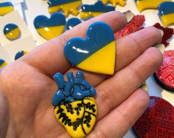 May include: A set of handmade pins featuring the Ukrainian flag in different shapes, including a heart, a human heart, and the outline of Ukraine. The pins are made of resin and have a glossy finish.
