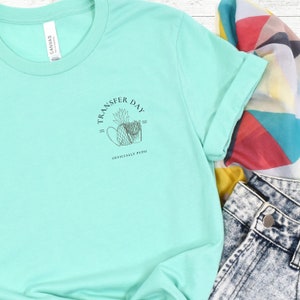 Puede incluir: Una camiseta turquesa con un gráfico negro de una piña y papas fritas con el texto "Transfer Day = Officially Pupo".