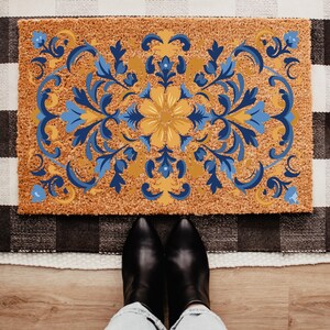 May include: A rectangular doormat with a floral design in shades of blue and yellow on a brown background. The mat is placed on a black and white checkered rug. Black boots and blue jeans are visible at the bottom of the image.