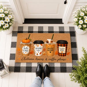 May include: A rectangular tan doormat featuring four coffee cups with autumn-themed designs and the text "hello FALL", "AUTUMN vibes", "SWEATER WEATHER", and "PUMPKIN SPICE". The mat includes the phrase "Autumn leaves & coffee please".