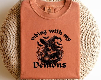 Op de afbeelding: Een roestoranje t-shirt met een zwarte grafische print van een heks met een puntmuts die een ketel vasthoudt, omringd door vleermuizen en takken. De tekst luidt "vibing with my Demons" in een gestileerd lettertype. Comfort Colors label.