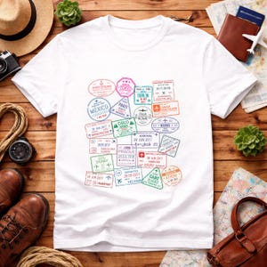 Puede incluir: Camiseta blanca con sellos de viaje coloridos de ciudades como Tokio, París y Roma. Los sellos incluyen fechas y códigos de aeropuerto, creando un tema de viaje. La camiseta se muestra sobre una superficie de madera.
