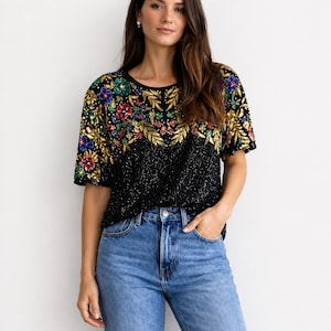 Peut inclure: Haut noir à sequins avec broderies florales dorées, rouges, bleues et vertes. Le haut à manches courtes a une encolure ronde et est associé à un jean en denim bleu clair. Le motif floral du haut est concentré sur la partie supérieure.