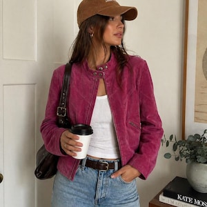 May include: A magenta suede jacket with a quilted design, paired with light-wash blue jeans and a brown baseball cap. The outfit includes a white tank top, a brown leather belt, and a dark brown shoulder bag.