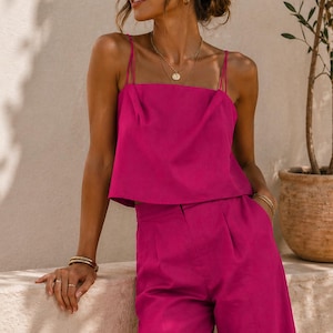 May include: A magenta two-piece outfit consisting of a square-neck crop top with spaghetti straps and high-waisted, wide-leg pants. The outfit is accessorized with gold jewelry and sunglasses. A potted plant is in the background.