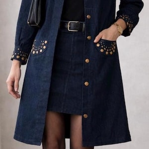May include: A dark blue denim coat dress with gold-colored stud embellishments on the collar, pockets, and cuffs. The dress features a matching denim skirt, black belt, and knee-high black boots. A black purse hangs from the shoulder.