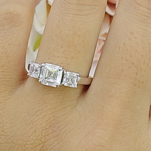 Könnte beinhalten: Ein silberner Ring mit drei quadratischen, klaren Edelsteinen. Der Ring befindet sich an einem Finger.