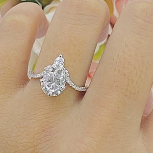 May include: A pear-shaped diamond engagement ring with a smaller round diamond on the side, set in a silver band. The ring is on a hand with a white background.