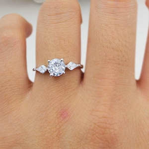 Puede incluir: Un anillo de plata con una piedra central redonda y dos piedras en forma de pera a cada lado.