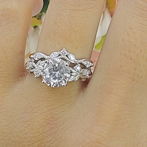 May include: A silver engagement ring set with a large, round, clear gemstone. The band features a floral design with smaller gemstones. The ring is on a person's finger, with a soft focus background of pink and white flowers.