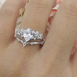 May include: A silver ring set with a pear-shaped clear stone surrounded by smaller clear stones. The ring is on a finger with a white background.