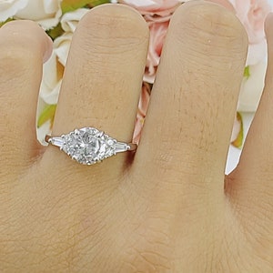 May include: A silver ring with a large round center stone and two smaller stones on either side. The ring is on a person's finger.