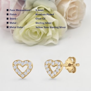 May include: A pair of gold heart-shaped earrings with clear cubic zirconia stones. The earrings have a hollow heart design and are 5.4 mm in size. The earrings are made of sterling silver with a yellow tone finish and rhodium plating.