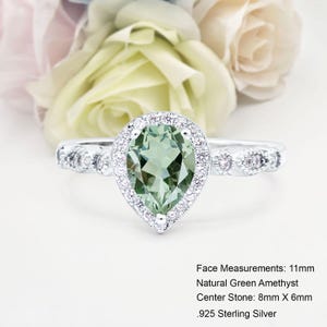 以下が含まれることがあります： 小さなダイヤモンドのハローで飾られた、ペアシェイプのグリーンアメジストがスターリングシルバーにセットされたリング。リングの幅は11mm、センターストーンは8mm×6mm。フェイスのサイズ：11mm 天然グリーンアメジスト センターストーン：8mm×6mm 925スターリングシルバー