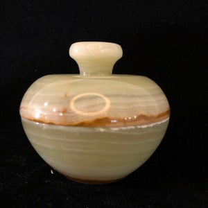 Peut inclure: Un petit pot décoratif rond avec un couvercle. Le pot est fait d'une pierre verte clair et marron. Le couvercle est d'une nuance de vert plus claire. Le pot a un corps bulbeux et un col court et cylindrique.