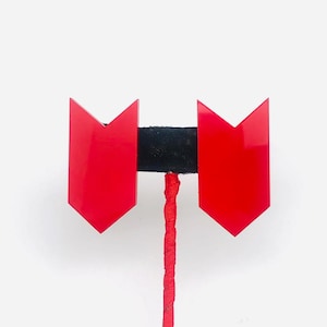 May include: A pair of red, arrow-shaped earrings. The earrings are connected by a black bar and a red, twisted cord. The earrings have a glossy finish and are set against a white background.
