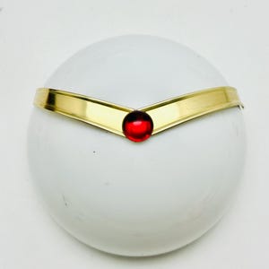 May include: A gold-colored metal tiara with a central, round, red jewel. The tiara is set against a white, rounded background, creating a striking contrast. The design is simple yet elegant, with a polished finish.