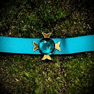 May include: A teal blue ribbon with a gold cross-shaped pin featuring a large, faceted teal blue gemstone.