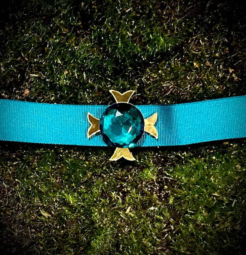 May include: A teal blue ribbon with a gold cross-shaped pin featuring a large, faceted teal blue gemstone.